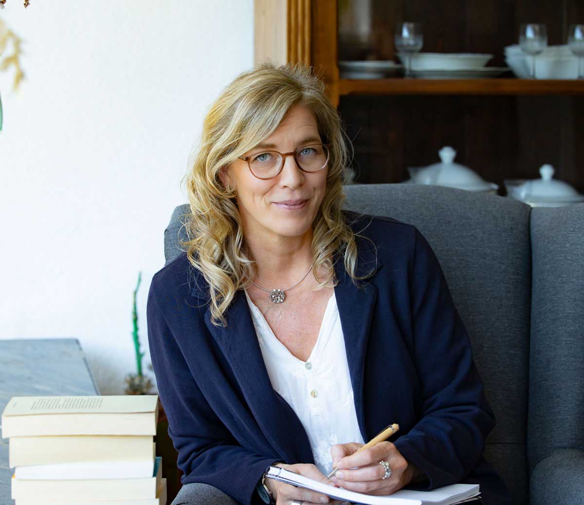 Frau mit blonden lockigen Haaren sitzt an einem Tisch mit mehreren Büchern und schreibt in ein Notizbuch